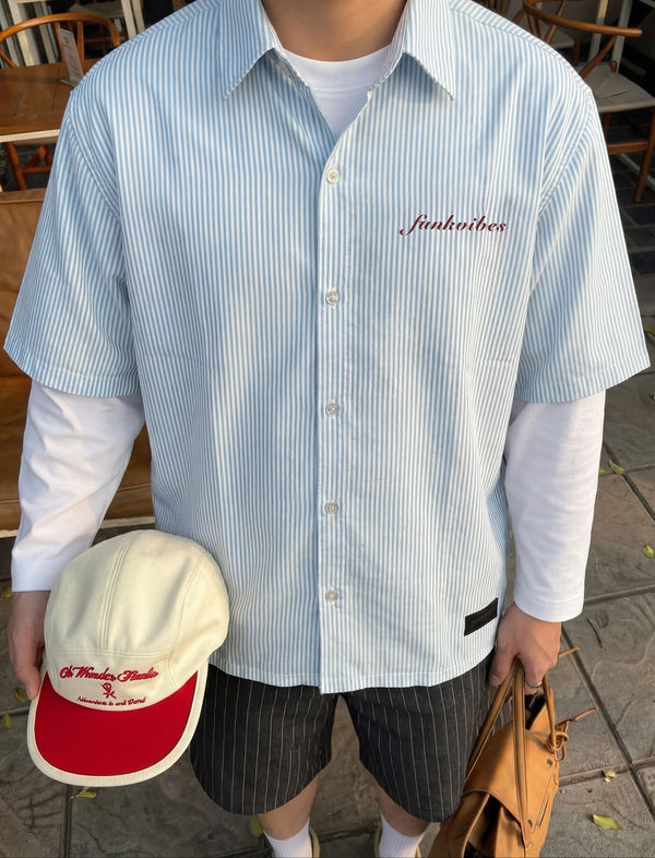 Person wearing a light blue striped shirt with 'Junkfood' logo, holding a cap and bag in an outdoor setting.