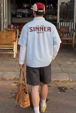 Person wearing a 'Sinner Society' shirt walking outside a building with chairs and tables.