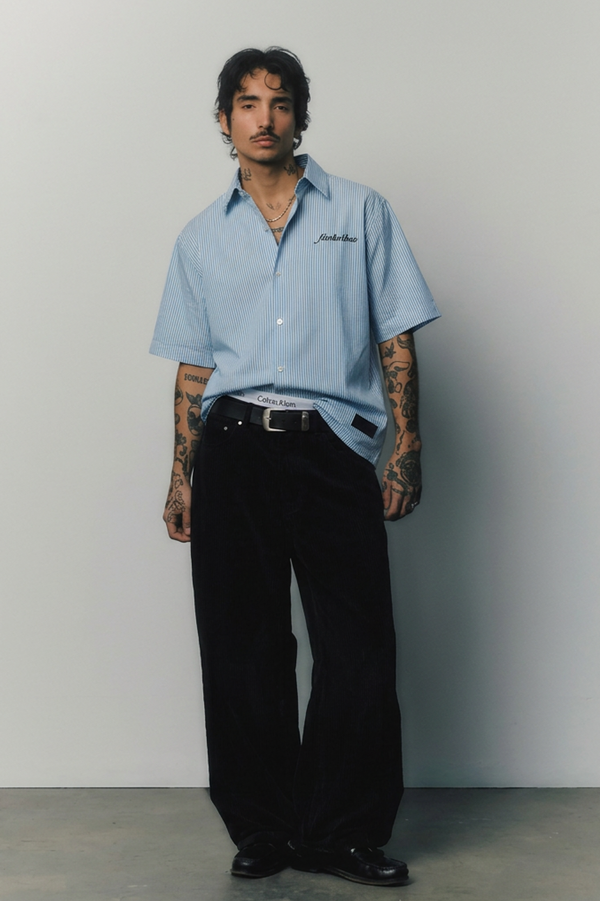 Person wearing a light blue shirt with a logo and black pants against a plain background