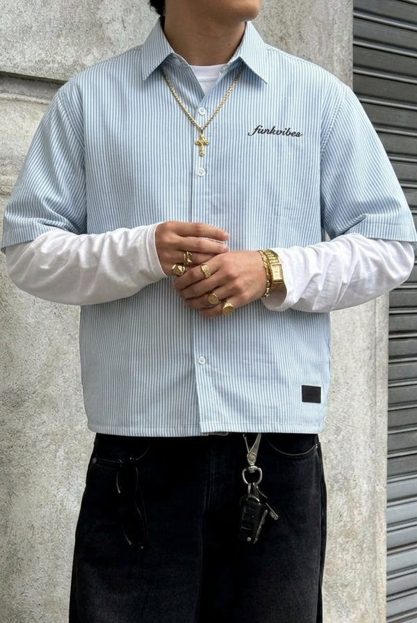 Person wearing a light blue striped shirt with a brand logo, standing against a concrete wall.