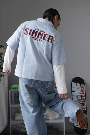 Person wearing a light blue shirt with 'Sinner Society' text, standing in a room with a plant and clothes rack.