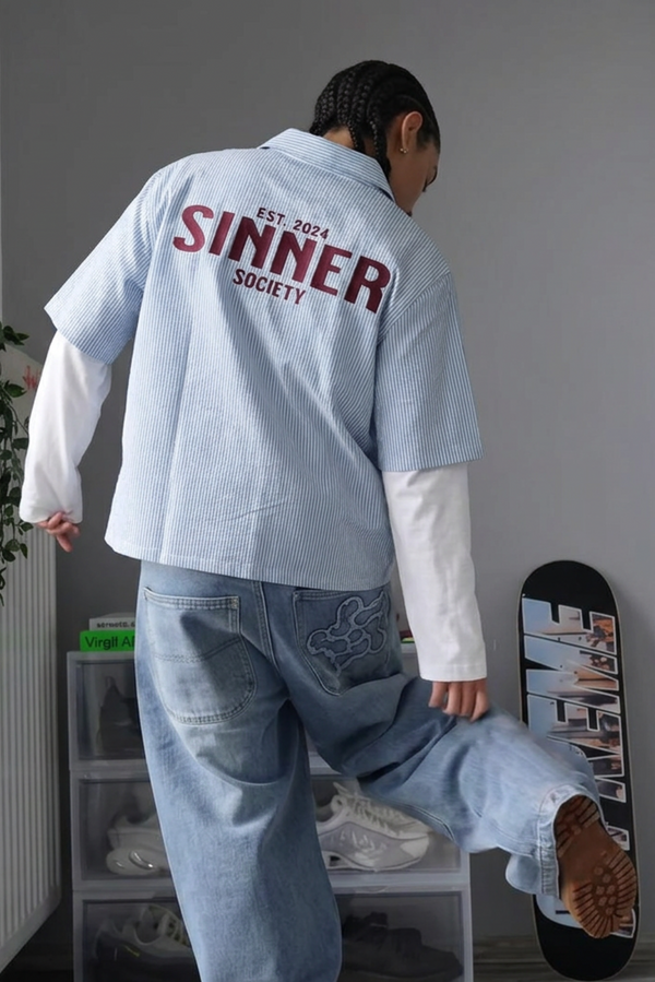Person wearing a light blue shirt with 'Sinner Society' text, standing in a room with a plant and clothes rack.